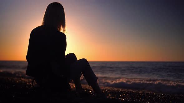 blonde in denim suit is sitting on seashore at sunset with smartphone in hands