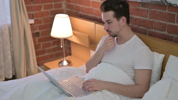 Young Man Thinking and Working on Laptop in Bed