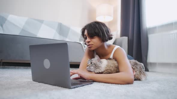 Young Female with a Cat Lies on the Floor in the Living Room and Works at a Laptop Selfisolation at