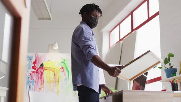 African american male artist wearing face mask holding a painting and using laptop at art studio