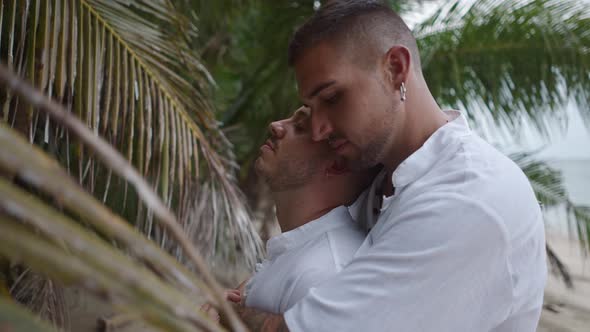 Gay Man Embracing His Partner From Behind Standing on a Tropical Beach By the Palm