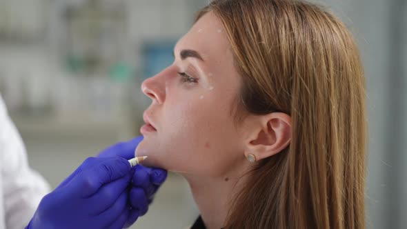 Headshot Side View of Slim Young Woman in Beauty Salon with Male Hand Marking Chin for Filler