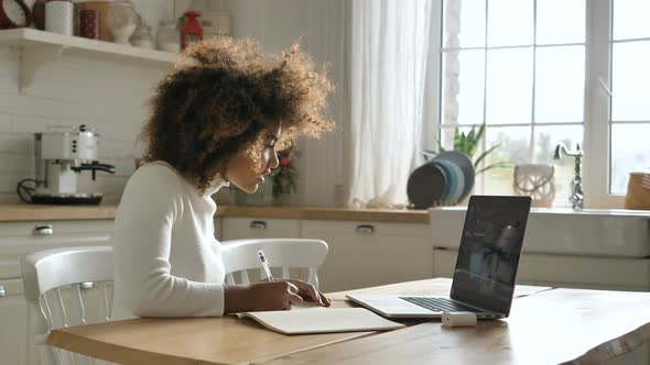 A Cheerful African-American Girl Listens and Takes Notes on an Online Lecture From Her Teacher