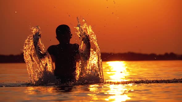 Silhouette of Boy at Sunset Raises Hands and Creating Splashes of Water. Slow Motion