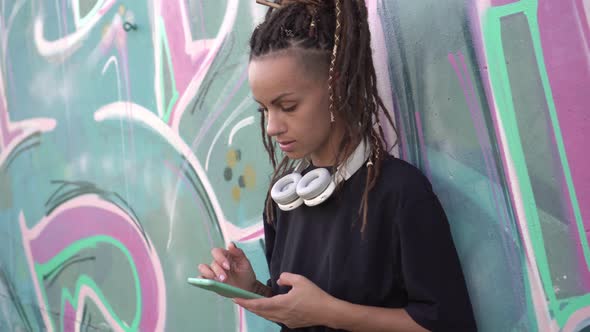 A Young Woman Looks Into a Smartphone Against the Background of a Wall with Graffiti Outdoors