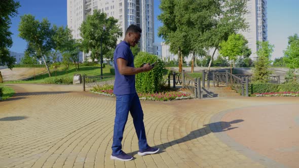 Smiling Afroamerican Man Walks Street Uses Mobile Phone Holding Cup with Coffee