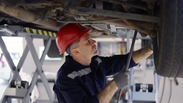 Auto Mechanic in Helmet Working Underneath Car Lifting Machine at the Garage