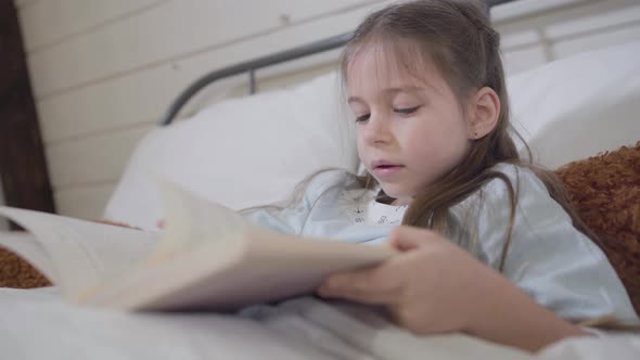 Close-up of Little Brunette Caucasian Girl Reading Book in Bed. Happy Charming Child Enjoying