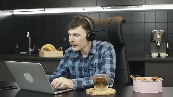 Young male freelancer using notebook sitting in kitchen at home listening to music in headphones
