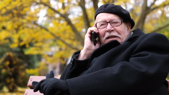 Positive Old Man Relax Autumn City October Park Sit on a Bench Call a Second Smartphone