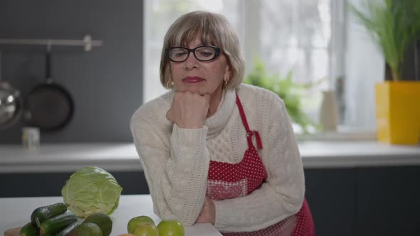 Portrait of Sad Beautiful Lonely Retiree in Eyeglasses Looking at Camera Standing at Kitchen