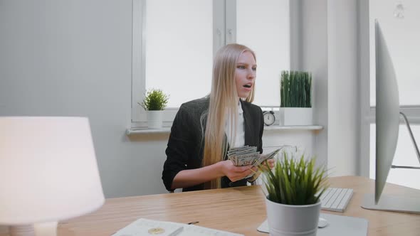 Woman Celebrating Success in Office. Elegant Blond Female Sitting at Workplace Holding in Hands