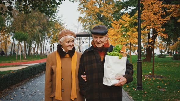 Aged Pair Talking and Smiling Walking in Autumn Park Holding Paper Bag of Groceries Baguette and