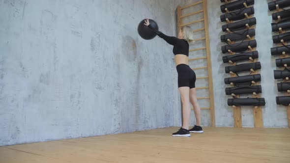 young woman doing squats while throwing a medicine ball up against a wall in a dark color