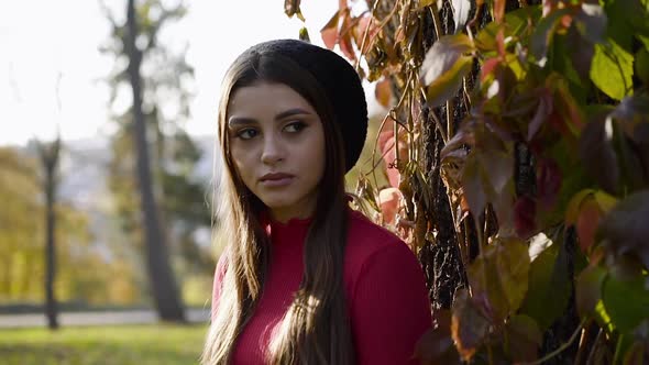 Attractive Longhaired Girl in a Cap Looking at Camera Near Autumn Tree Trunk