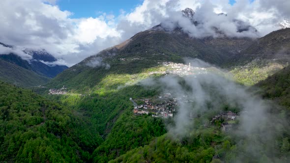 Clouds forming above scenic village in lush Italian alp valley hillside; drone