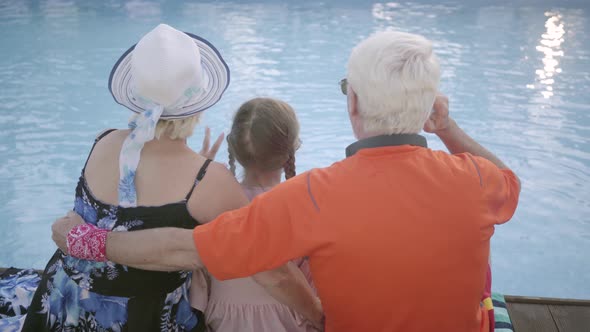 Mature Man, Woman and Little Girl Sitting on the Edge of the Pool, Back View