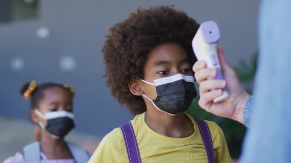 Diverse male teacher measuring temperature of schoolchildren, all wearing face masks