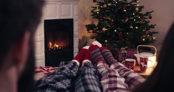 Over shoulder couple feet in christmas woolen socks near fireplace decorated tree tee cup in backg