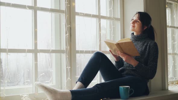 Dreamy Young Woman Is Reading Book and Smiling on Christmas Sitting on Window-sill Decorated with