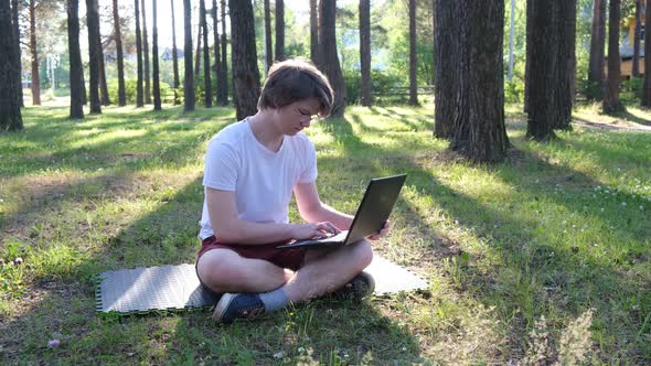 Young Man Sitting on the Grass in Park Working on His Laptop