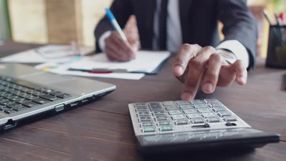 Black man office worker in a suit counts on calculator and records the results with a pen in paper