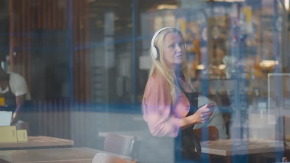 View Through Window of Cheerful Senior Waitress Listening To Music in Headphones and Dancing in Cafe