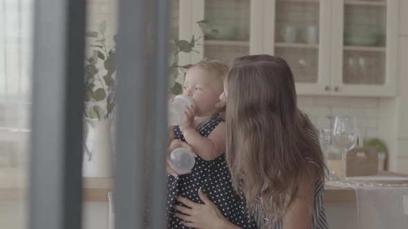 Beautiful Smiling Young Woman Showing Her Baby Girl How To Drink From the Baby Bottle in the Kitchen