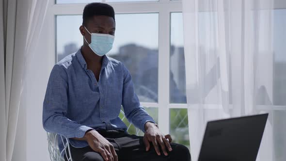 Young Man in Coronavirus Face Mask Sitting in Sunny Home Office Conferencing Online on Laptop