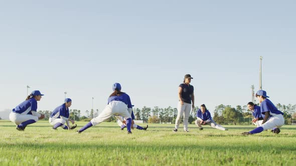 Diverse group of female baseball players and coach warming up on pitch, squatting, stretching legs