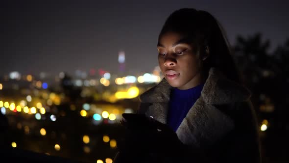 A Young Black Woman Works on a Smartphone and Smiles at the Camera in an Urban Area at Night