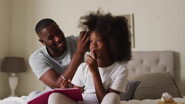 African american father tying his daughters hair while sitting on the bed at home