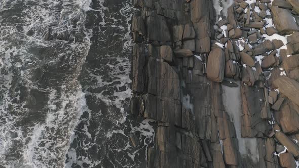 Flying slowly forward as waves lap the rocky shore, winter sunrise AERIAL TOP DOWN