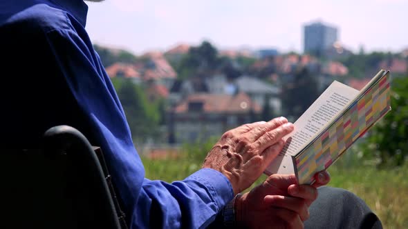 Old Senior Man Sits on the Bench in the Park and Reads Book - City in Background - Closeup