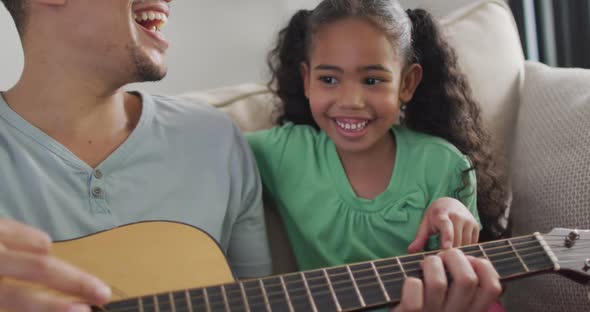 Happy biracial father and daughter sitting on sofa playing guitar