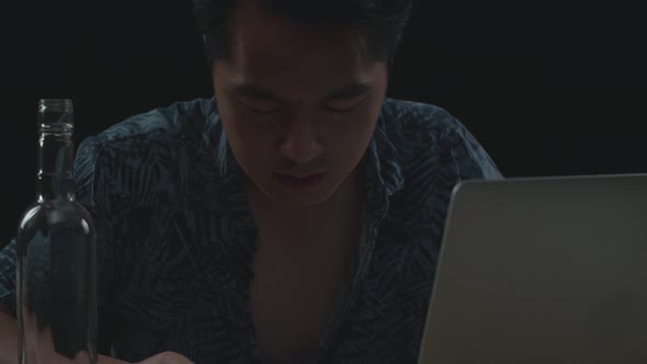 Close Up Of Asian Man Drinking Vodka While Working On A Laptop In Black Background