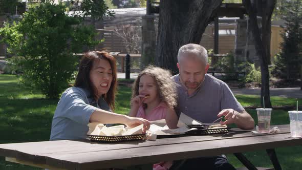 Family sitting at picnic table in park laughing and eating