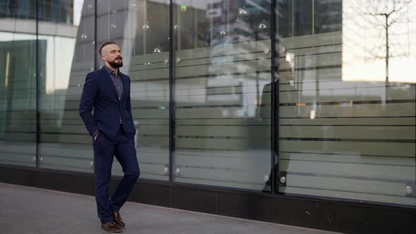 a Moustached and Bearded Man in a Blue Suit Has Hands in Pockets and is Walking Along the Glass Wall