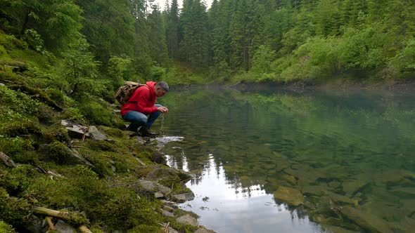 A Bearded Man with a Backpack Drinks Water From a Clean Lake