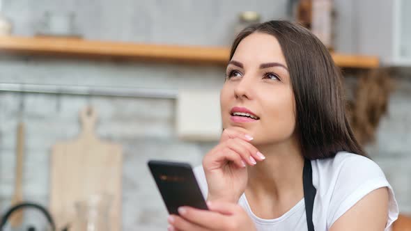 Beautiful Female Housewife in Apron Dreaming Chatting Using Smartphone at Modern Kitchen