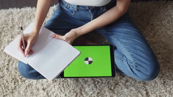 Woman is Sitting on Floor and Making Notes in Notebook Tablet with Green Screen is Lying Next to It