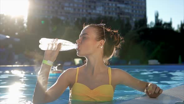 Young Woman in a Swimsuit Drinks Cool Water From a Plastic Bottle in the Pool