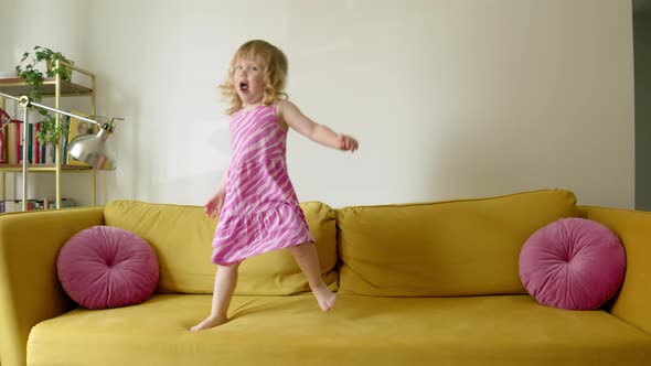 Cheerful and Cute Preschool Girl Jumps and Dances on Yellow Large Sofa in Living Room