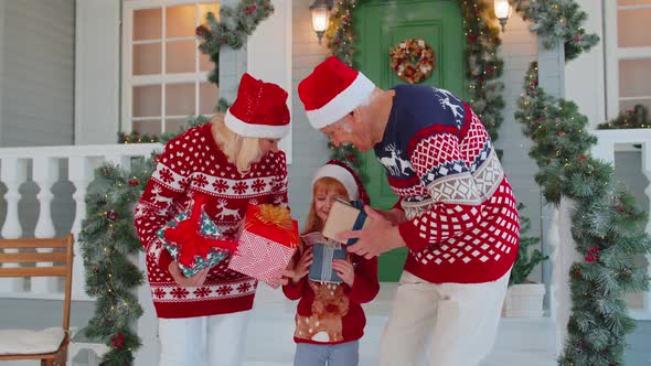 Senior Grandparents with Grandchild Girl Kid Exhcanging Gifts Near Christmas House Celebrating Xmas