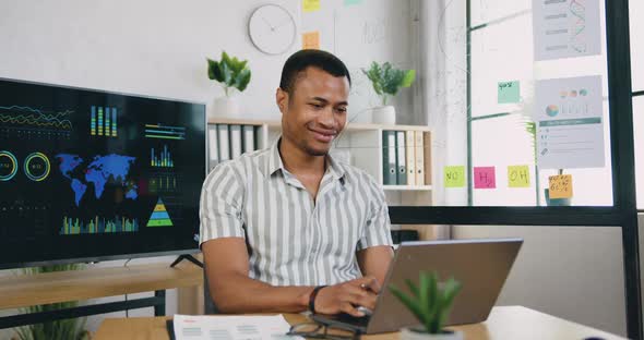 African American Office Worker Writing Out Important Notes Into Report while Working with Computer