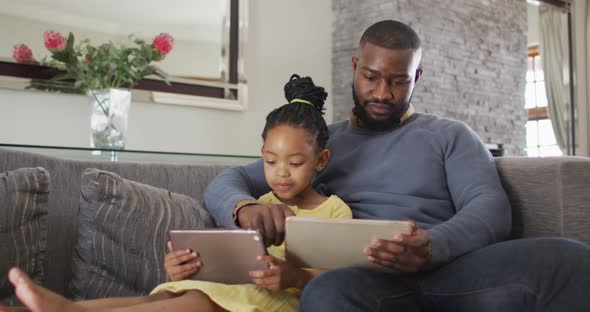 Happy african american daughter and father using tablets on sofa