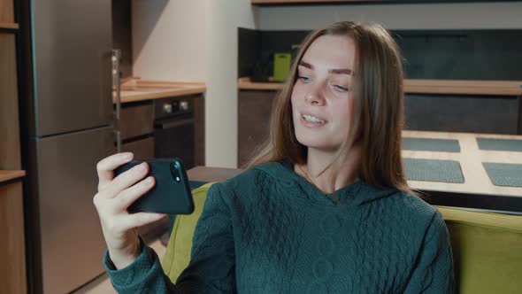 Smiling Relaxed Young Woman Hold Smartphone Watching Social Media Stories Video Siting on Sofa in