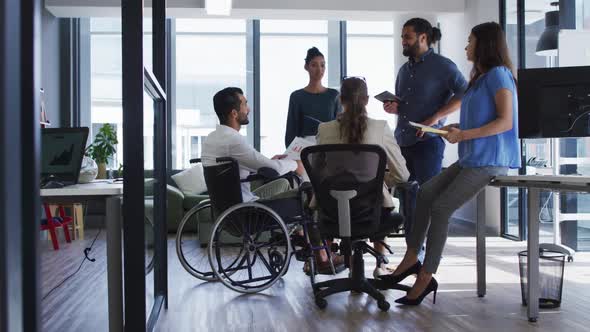 Diverse group of work colleagues talking at casual office meeting, one in wheelchair