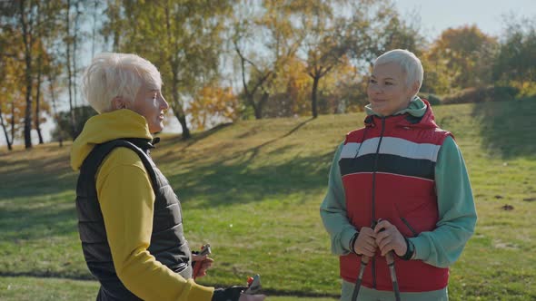 Two Aged Women Talk in Sunny Fall Park Wearing Vests and Holding Walking Poles