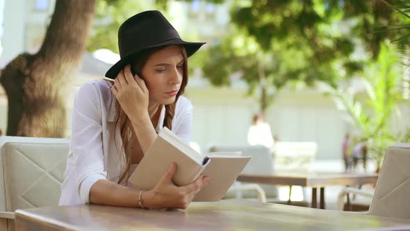 Young Beautiful Girl in Hat Reading Book in Cafe